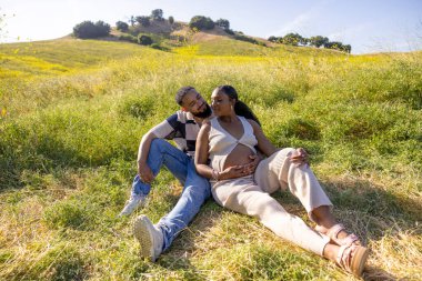 Neşeli bir Afrikalı Amerikalı çift doğada dinleniyor, hamileliği ve açık havanın güzelliğini manzaralı bir arka planla kutluyor.