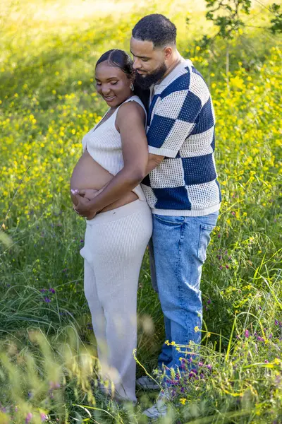 Afrika kökenli Amerikalı bir çift, açık havada huzurlu bir yaz gününün tadını çıkarırken, eşi, yeşil ve canlı çiçeklerin arasında, hamile karısına şefkatle sarılıyor.