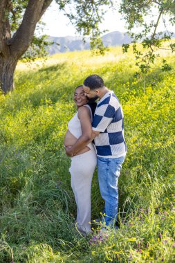 Güzel bir yaz gününde, sevecen bir Afrikalı Amerikalı çift açık havada, eşlerinin karnını nazikçe kucaklıyor, canlı yeşillik ve görkemli doğayla çevrili.
