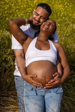 Sakin bir açık hava ortamında, neşeli bir Afrikalı Amerikalı çift hamileliklerini kutluyor, koca karısına sarılıyor, büyüyen aileleri için mutluluk saçıyor.