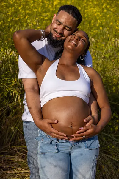 Sakin bir açık hava ortamında, neşeli bir Afrikalı Amerikalı çift hamileliklerini kutluyor, koca karısına sarılıyor, büyüyen aileleri için mutluluk saçıyor.
