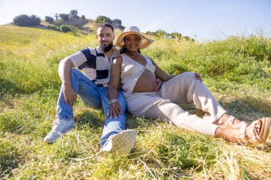 Neşeli bir Afrikalı Amerikalı çift yaz güneşinin altında dışarıda dinlenirken, hamileliğin güzel yolculuğunu kucaklarken ve çevresindeki manzarayı ve doğayı takdir ederken görülmektedir.