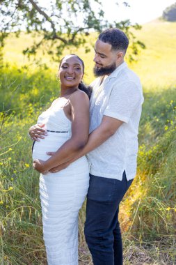 Yazın güzel bir açık hava ortamında neşeli bir Afrikalı Amerikalı çift hamileliklerini kutluyor. Koca, karısına şefkatle sarılıyor. Bebek çarpması, sevgi ve bağlılık yayıyor.