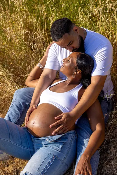 Neşeli hamile bir çift, dışarıda bir yaz gününün tadını çıkarırken, koca, eşinin karnına şefkatle sarılıyor, yaklaşan ebeveynliklerini kutluyor..