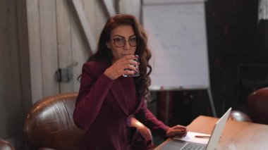 Modern bir ofiste, organize masasındaki kendine güvenen bir iş kadını dizüstü bilgisayarı üzerinde ustalıkla çalışırken bir içki içerek profesyonelliğini ve kariyerine olan bağlılığını gösteriyor.