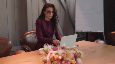 Güzel dekore edilmiş bir toplantı odasında şık bir takım elbise giymiş kendine güvenen bir iş kadını dikkatle dizüstü bilgisayarına odaklanarak işine ve profesyonel amaçlarına bağlılığını gösteriyor.