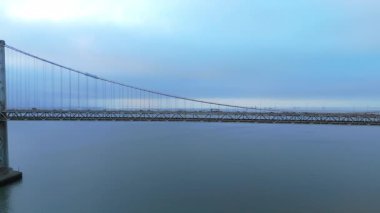 Büyüleyici panoramik dron görüntüleri San Francisco 'daki ikonik Bay Köprüsü' nü güzel bir şekilde gösteriyor. Sabah erken saatlerde sakin ve pitoresk bir su zeminine doğru hareket eden yoğun trafiği vurguluyor.