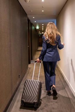 Zarif bir donanma elbisesi giymiş şık ve şık bir iş kadını bavullarını modern, aydınlık bir otel koridorunda incelikle taşıyarak profesyonel seyahat yaklaşımını etkili bir şekilde sergiledi.