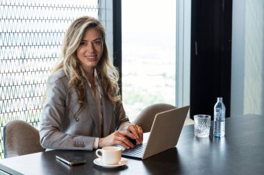 Modern bir ofiste, kendine güvenen bir iş kadını dizüstü bilgisayarıyla çalışır, çarpıcı bir şehir manzarasının keyfini çıkararak, profesyonelliğini, kararlılığını ve görevlerine yoğun bir şekilde odaklanmasını vurgular.