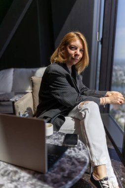 Tarz sahibi genç bir kadın, modern ofis ortamında güvenle oturur, kararlılıkla pencereden dışarı bakar, günümüzün üretkenlik ve modern iş kültürünün özünü bütünüyle somutlaştırır.