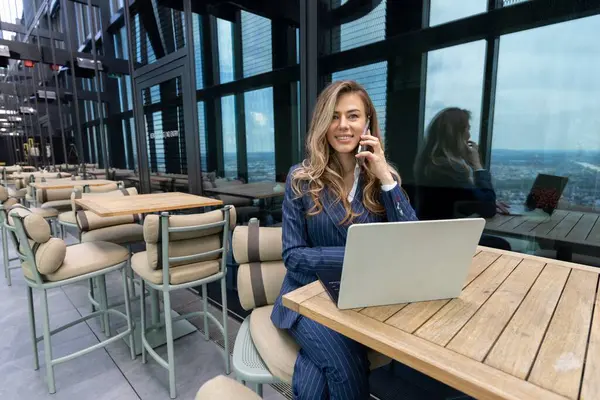 Profesyonel takım elbiseli kendine güvenen bir kadın önemli bir iş görüşmesinde. Şaşaalı şehir manzaralı, rahatlama ve üretkenliği dengeleyen bir çatı katında dizüstü bilgisayarını ustalıkla kullanıyor.