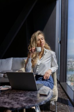 Şık bir iş kadını, şık bir ofiste dizüstü bilgisayarıyla çalışırken kahveden zevk alır. Şehir manzarası, zarafet ve verimliliği harmanlayarak modern girişimciliği somutlaştırır.