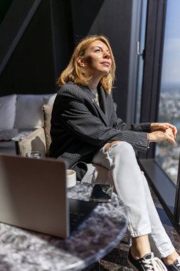 Spor ceketli şık bir kadın modern bir ofiste büyük bir pencerenin yanında oturuyor ve şehir manzarasının tadını çıkarıyor. Bu sahnede modern bir iş kadınının iş hayatı, verimliliği ve şık yaşam tarzı vurgulanıyor.