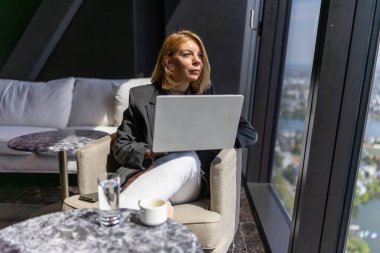 Modern bir ofiste, odaklanmış bir kadın laptopuyla çalışır, büyük pencerelerden çarpıcı şehir manzarasının tadını çıkararak, görevlerini yaratıcı bir şekilde yaparken üretkenliği ve ilhamı somutlaştırır.