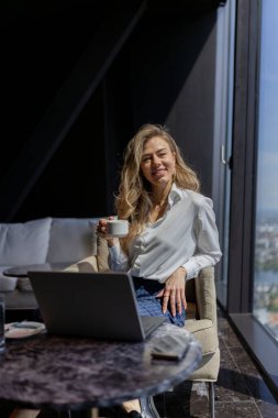 Güzel kıvırcık saçları olan kendine güvenen bir kadın, şehrin çarpıcı panoramik manzarasını barındıran modaya uygun ve şık bir ofis ortamında laptopuyla çalışırken kahvesini yudumlamayı seviyor.