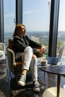 Şık bir genç kadın, elinde bir dizüstü bilgisayar ve bir fincan sıcak kahveyle dışarıdaki şehrin çarpıcı manzarasına hayran kalarak eğleniyor.