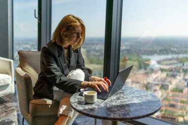 Odaklanmış ve kararlı bir iş kadını, iş hayatındaki üretkenlik ve modern çalışma alanlarını somutlaştırarak kendisine ilham veren çarpıcı bir ufuk çizgisi manzarasının keyfini çıkarırken bilgisayarından özenle faydalanıyor.