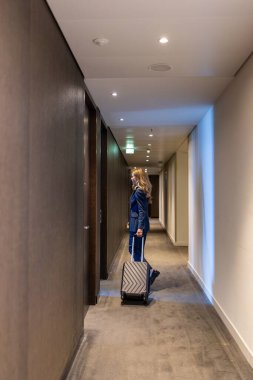 Zarif, mavi bir elbise giymiş bir gezgin, uzun, modern otel koridorunda pürüzsüz bir bavul yuvarlar, her adımda seyahat, keşif ve maceranın özünü yakalar.