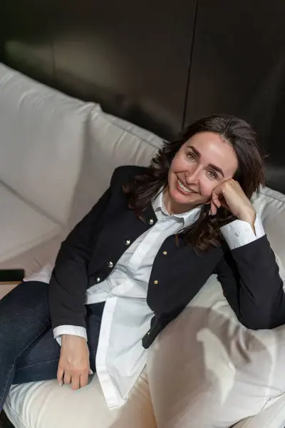 Şık ve sofistike bir kıyafet giymiş kendine güvenen genç bir kadın şık, rahat bir koltukta zarif bir şekilde oturur. Şık, modern bir iç mekanda sıcaklık ve zarafet saçar.