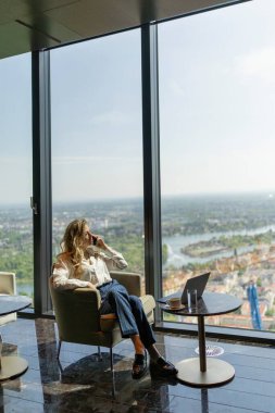 Şık giyimli bir kadın lüks, çağdaş bir mekanda oturur, yüksek apartman dairesinin geniş şehir manzarasını düşünür, hırs, hırs ve başarı hayalleriyle dolu.