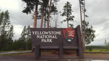 İkonik Yellowstone Ulusal Parkı giriş tabelasının çarpıcı manzarası gür ağaçlar ve dramatik bir gökyüzü tarafından çerçevelenmiştir. Bu da izleyicilerde güçlü bir macera ve keşif hissi uyandırır.