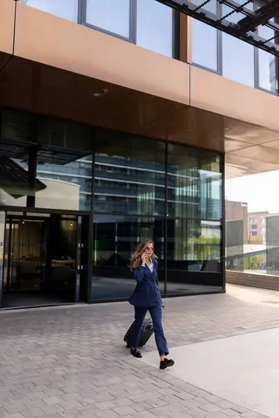 Şık bir takım elbise giyen kararlı profesyonel bir kadın şık, çağdaş bir ofis binasının dışında güvenle yürür. Bu da canlı ve başarılı bir iş ortamının özünü yansıtır.