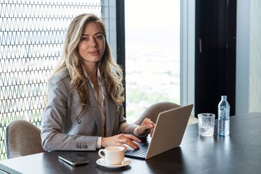 Çarpıcı şehir manzaralı modern bir ofiste, kendine güveni tam bir kadın laptopuyla çalışıyor, yakındaki dumanı tüten bir fincan kahve, şık çalışma alanındaki verimliliği ve motivasyonu sembolize ediyor.