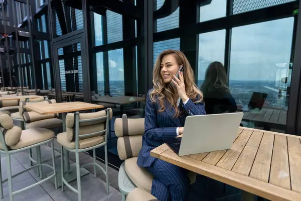 Kendine güvenen bir iş kadını, panoramik manzarası olan modern bir çatı katındaki bir kafede laptopuyla çalışırken bir telefon görüşmesi yaptı.