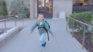 Aydınlık ve güneşli bir günde, renkli bir sırt çantası takan neşeli küçük bir çocuk heyecanla okulun girişine doğru yürüyor, öğrenme ve eğlenceli aktivitelerle dolu bir günü kucaklamaya hazır.