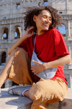 Şık bir genç, parlak kırmızı bir tişört giyerek mutlu bir şekilde ışıldıyor. Rahat bej bir şortla, ünlü Colosseum 'un yanına oturmuş, neşeli ve canlı bir anı yakalıyor.