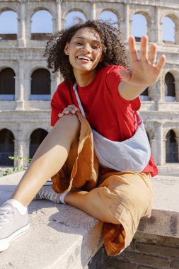 Neşeli genç bir kadın taş bir çıkıntıda otururken kameraya el sallıyor, arka planda Colosseum, Roma 'daki gezi macerası sırasında canlı kişiliğini sergiliyor.