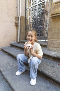 Büyüleyici bir şehir ortamında, neşeli küçük bir çocuk merdivenlerde oturur, lezzetli bir aperatifin üzerinde mutlu bir şekilde kıtır kıtır yemek yer, ifadeleri ılık gün ışığında saf zevk ve masumiyet yayar.