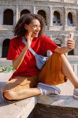 Günlük kıyafetlerle neşeli genç bir kadın taş bir çıkıntıda oturur, Roma 'daki ikonik Colosseum' un önünde selfie çekerken el sallar, seyahat macerasının harika bir anını yakalar.