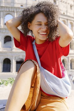Güzel kıvırcık saçlı neşeli genç bir kadın Roma 'daki ikonik Colosseum' da mutlu bir şekilde poz veriyor. Parlak güneşli bir günün tadını çıkarırken canlı bir kıyafet sergiliyor. Mutluluk ve pozitiflik saçıyor.