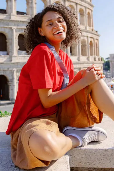 Canlı kırmızı gömlekli ve şık kahverengi etekli neşeli genç bir kadın muhteşem Colosseum 'un önünde mutlu bir şekilde oturur. Seyahatin ve unutulmaz maceranın özünü kucaklar.