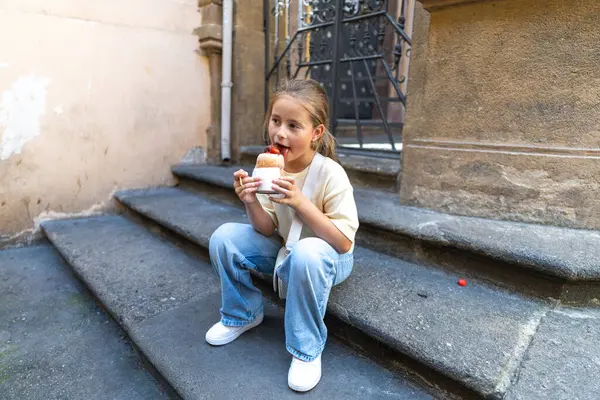 Neşeli bir çocuk sıcak taş basamaklarda oturuyor, canlı bir şehir ortamında lezzetli bir tatlının tadını çıkarıyor. Bu sahne, çocukluk deneyimlerinin neşe ve basitliğini yakalar ve hayata neşe katar.