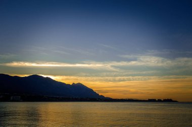 Gün batımını deniz üzerinde parlak gökyüzü solda bulutlarla görebilirsiniz dağlar artık güneş tarafından aydınlatılmıyor ve oryantasyon manzara için bir yer var yazılar için bir yer.