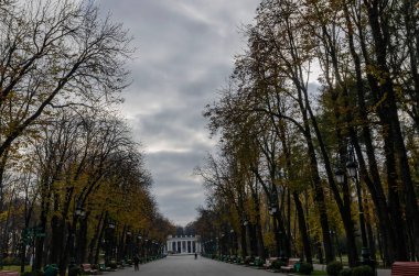 Bulutlu bir gökyüzüne karşı beyaz bir binaya açılan bir ara sokağa sahip sonbahar parkı.
