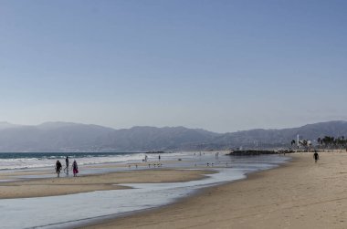 Venice Beach, California, America People walk along the beach at sunset, reflected in the water, rolled by the waves.