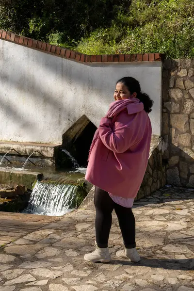 Pembe paltolu bir kadın taş bir duvarın önünde duruyor. Gülümsüyor ve manzaranın tadını çıkarıyor.