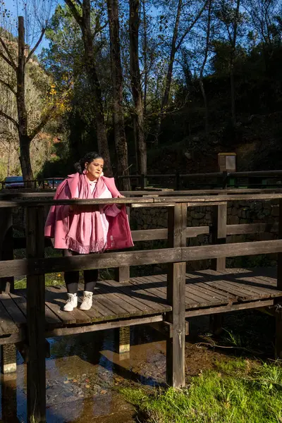 Pembe paltolu bir kadın ahşap bir köprüde duruyor ve bir su kütlesine bakıyor. Sahne huzurlu ve sakin, kadının bakışları suya çevrilmiş.