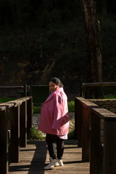 Pembe paltolu bir kadın ahşap bir köprüde duruyor. Gülümsüyor ve kameraya bakıyor.