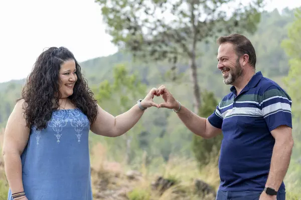 Bir erkek ve bir kadın el ele tutuşup parmaklarıyla kalp şekli oluşturuyorlar. Adam mavi bir gömlek giyiyor ve kadın mavi bir elbise giyiyor. Sahne mutlu ve romantik.