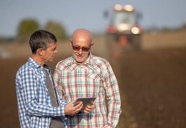 İki kıdemli çiftçi, sonbaharda tarlada traktörle tarla işlerken tablete bakıyor.