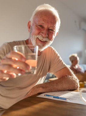 Happy senior man sitting at dining table at home holding glass of water and cheers. Healthy lifestyle in elderly time concept