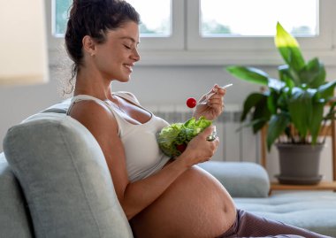 Evdeki kanepede salata yiyen güzel genç bir kadın. Annelik dönemi boyunca sağlıklı yaşam biçimi
