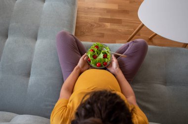 Hamile kadın kanepede oturuyor ve elinde salatayla kaseyi tutuyor. Annelik dönemindeki sağlıklı yaşam tarzı