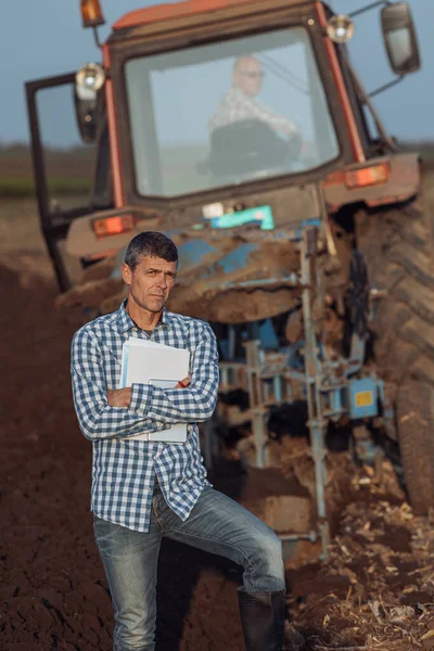 Mature farmer with notebook and tablet standing in front of tractor hoeing in field in autumn