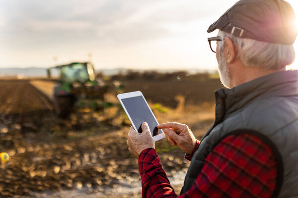 Серьезный зрелый фермер смотрит на планшет перед трактором в поле осенью