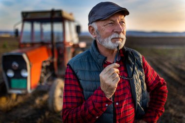 Pensive mature farmer standing in front of tractor in field in autumn